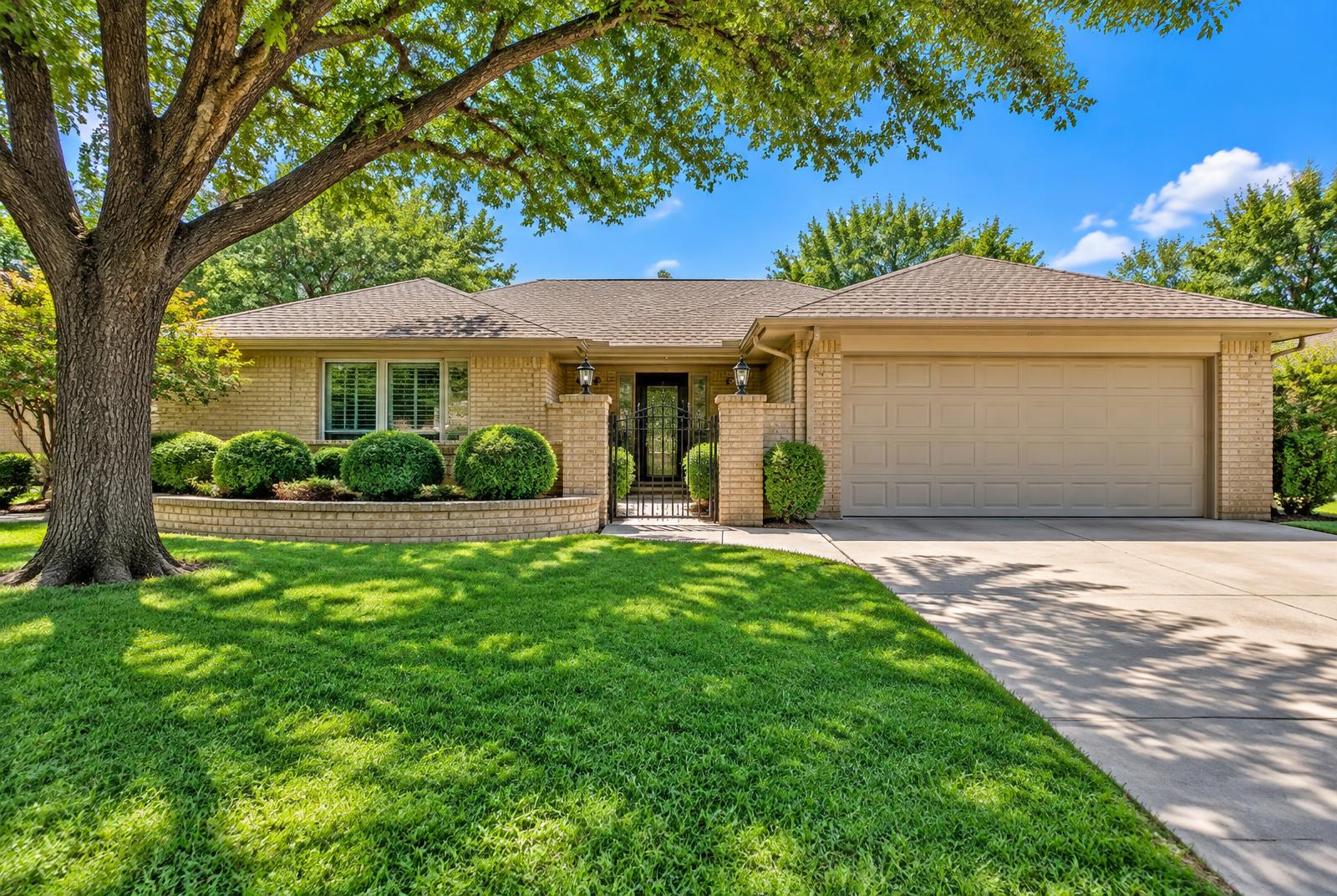 Front of the tan brick home with shade tree, wrought-iron entry gate, and manicured shrubs